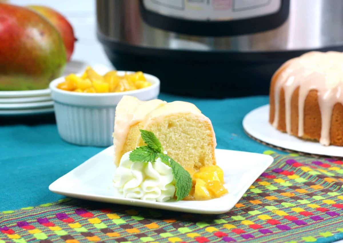 A slice of glazed bundt cake on a white plate with whipped cream, mint, and diced mango; a whole bundt cake, mango, and ramekin in the background.