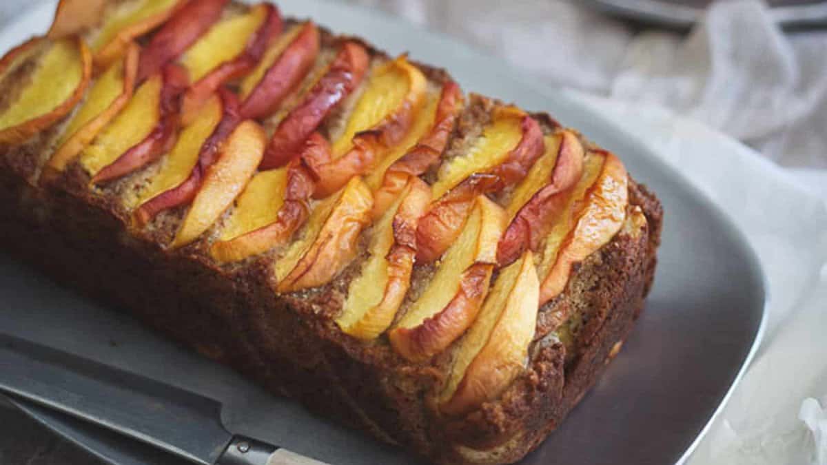 A loaf of bread or cake topped with neatly arranged slices of peaches sits on a gray plate next to a knife.