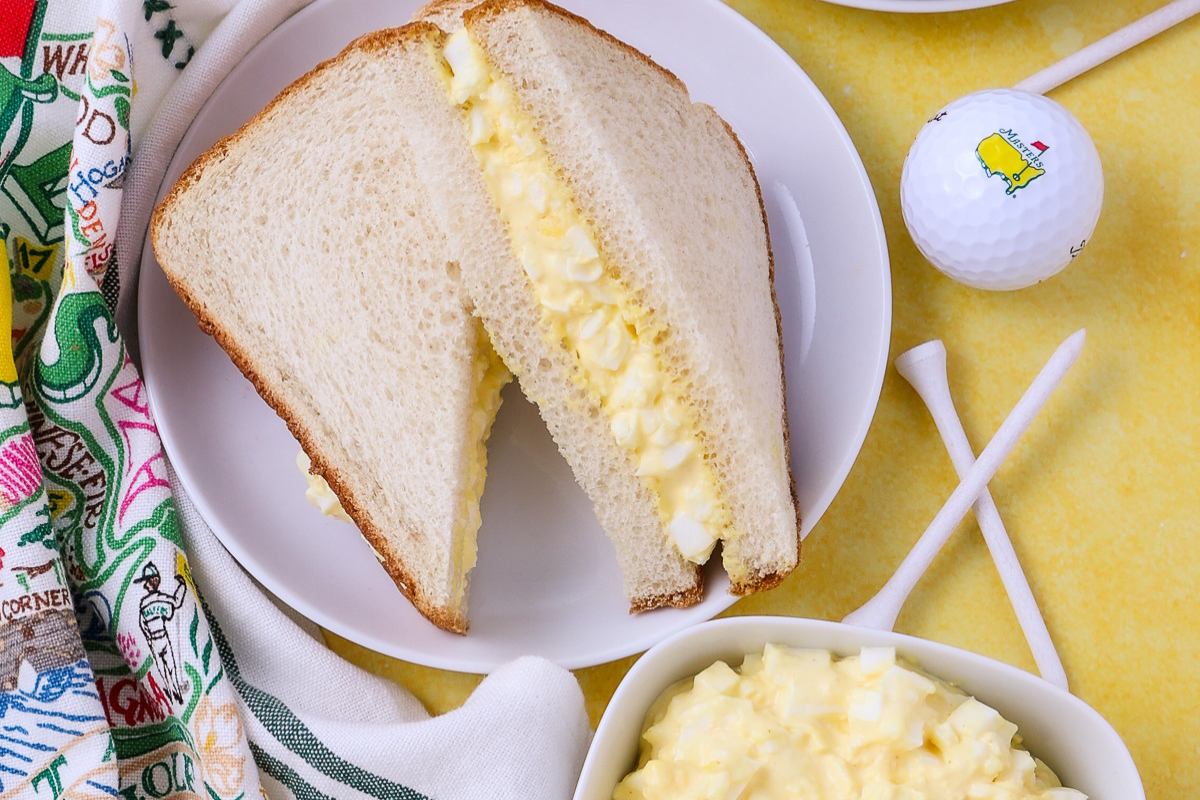 A plate with an egg salad sandwich cut in half, placed next to a golf ball, tees, and a patterned cloth on a yellow surface.