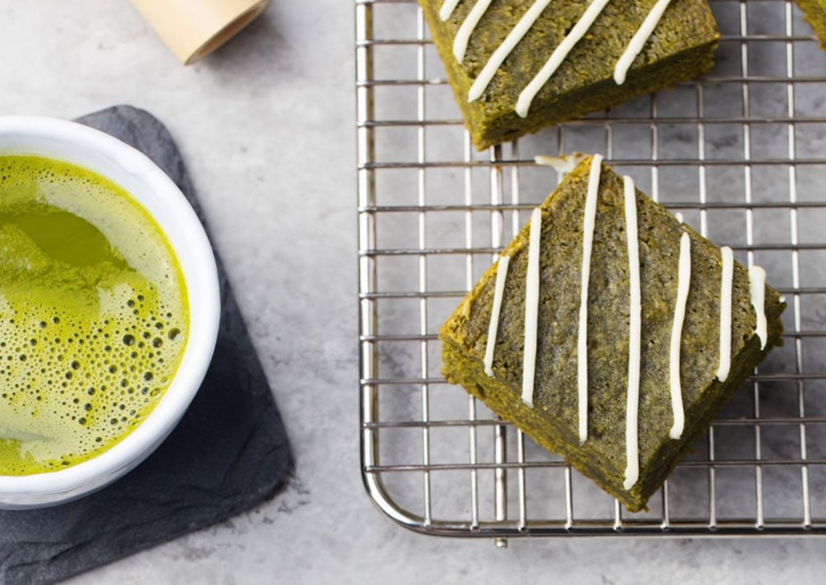 A cup of matcha tea sits beside two matcha brownies with white icing on a wire rack, on a light stone surface.