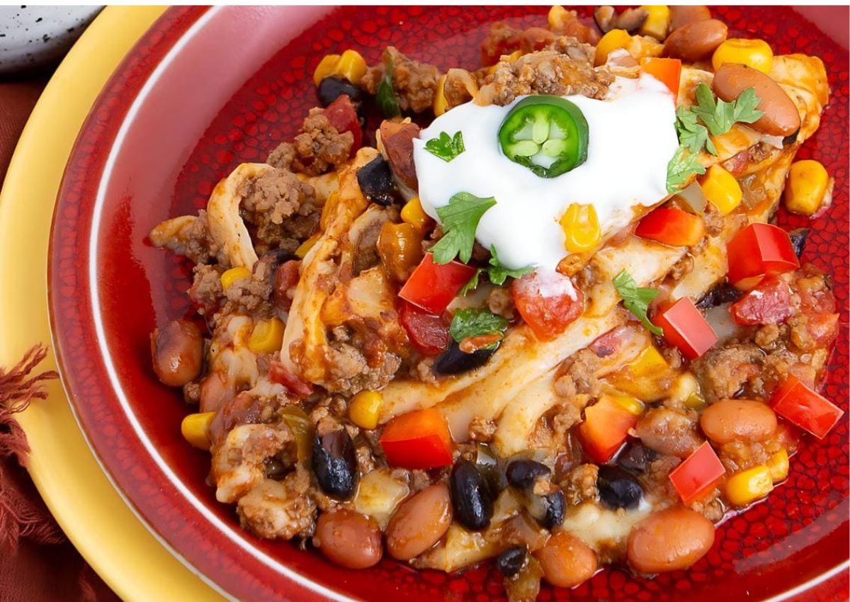 A plate of Mexican casserole with ground beef, beans, corn, tomatoes, and cheese, topped with sour cream, cilantro, diced red pepper, and a jalapeรฑo slice.