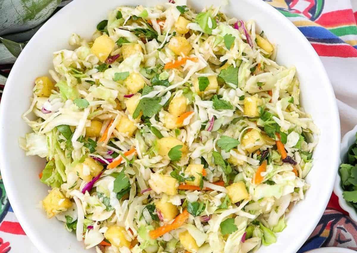 A bowl of coleslaw with shredded cabbage, carrots, cilantro, and pieces of pineapple on a colorful tablecloth.