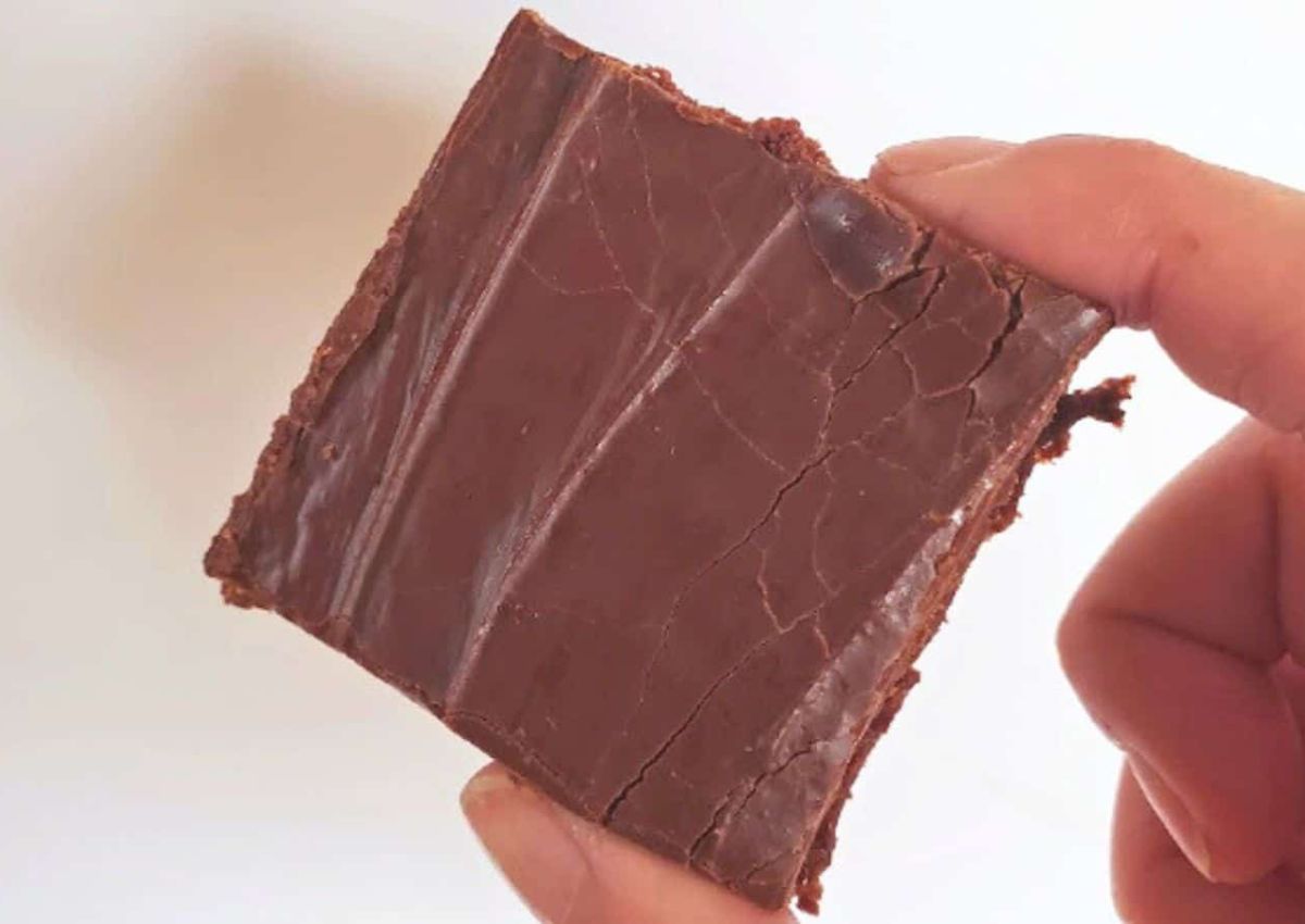 A hand holding a square piece of chocolate fudge with visible cracks on the surface.
