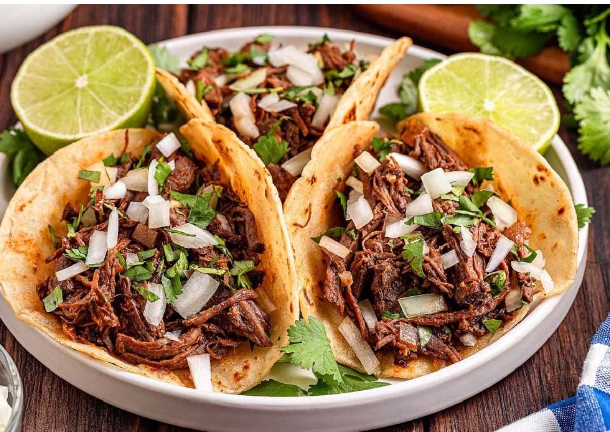 Three beef tacos topped with chopped onions and cilantro are served on a plate with lime halves and fresh cilantro garnish in the background.