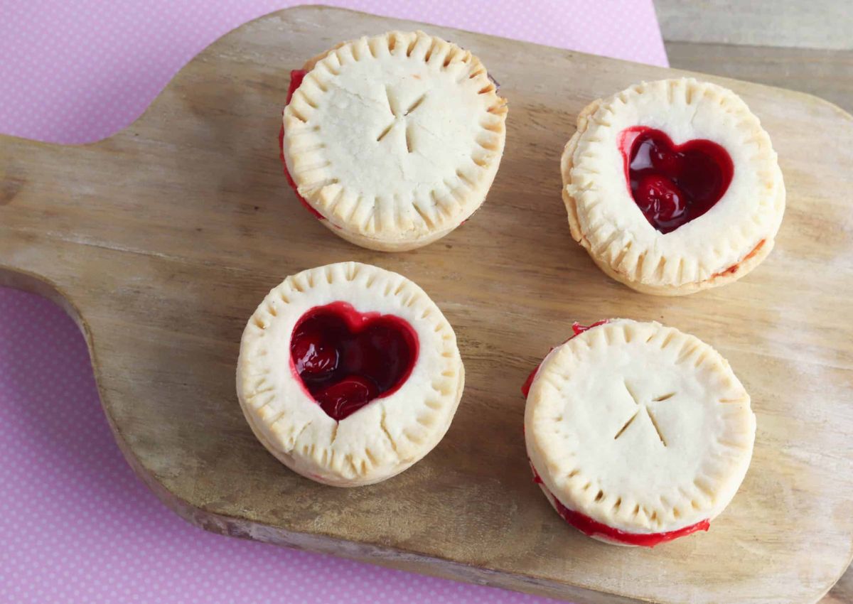 Four small cherry pies on a wooden board; two have heart-shaped cutouts showing the filling, while the other two have solid tops with an X-shaped vent.