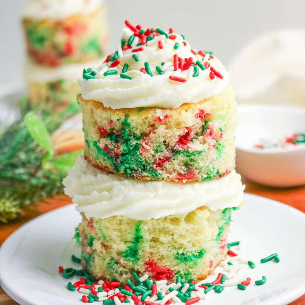 A stack of two mini vanilla cakes with red and green sprinkles, topped with white frosting and more sprinkles, on a white plate.
