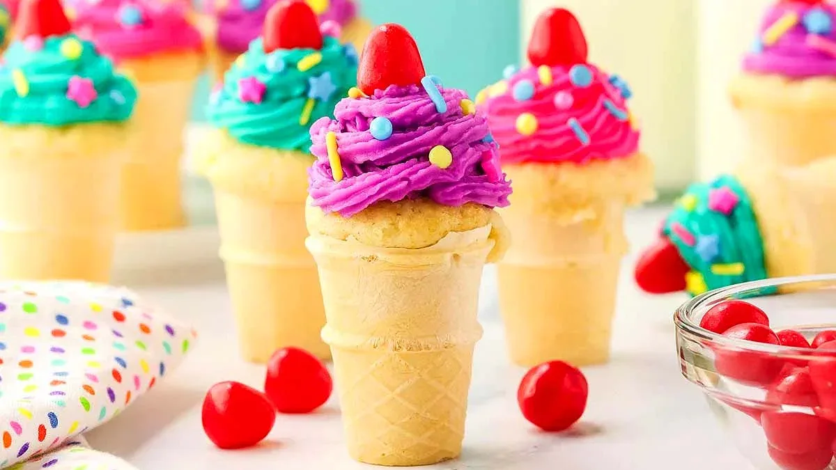 Ice cream cone cupcakes with colorful frosting and sprinkles, topped with a red candy, arranged on a table with additional candies nearby.