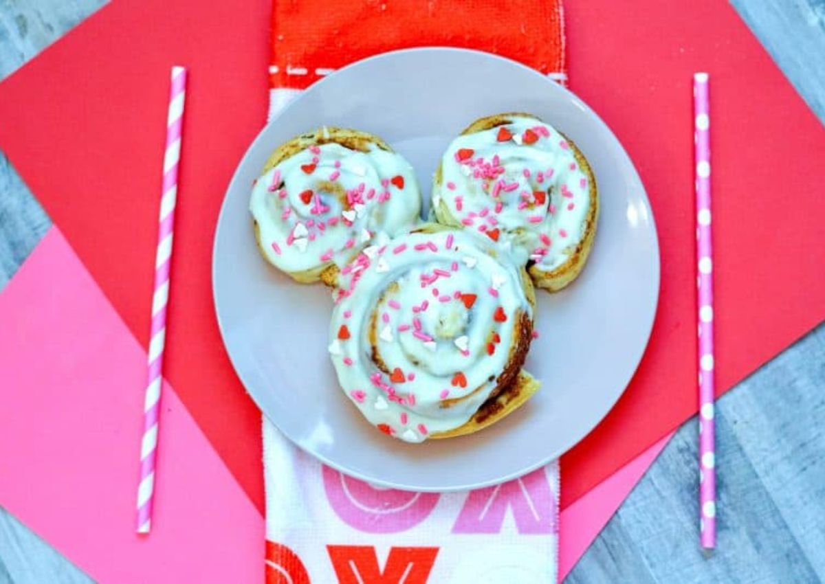 Three cinnamon rolls with white icing and pink heart sprinkles arranged in a Mickey Mouse shape on a white plate, with pink straws and red decorations.