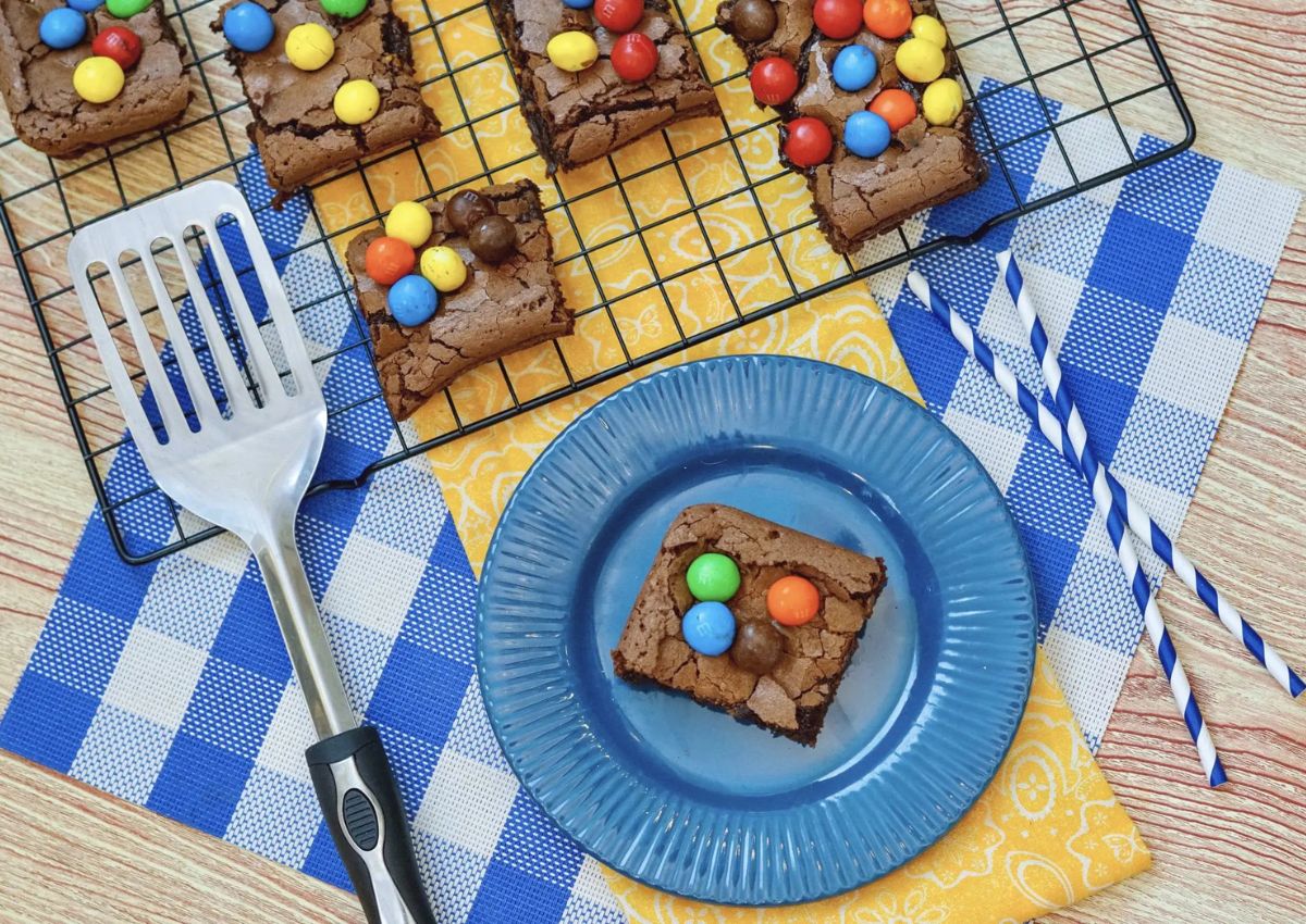 Brownies topped with colorful candy-coated chocolates on a cooling rack and blue plate, with a spatula, yellow napkin, blue straws, and blue checkered tablecloth.