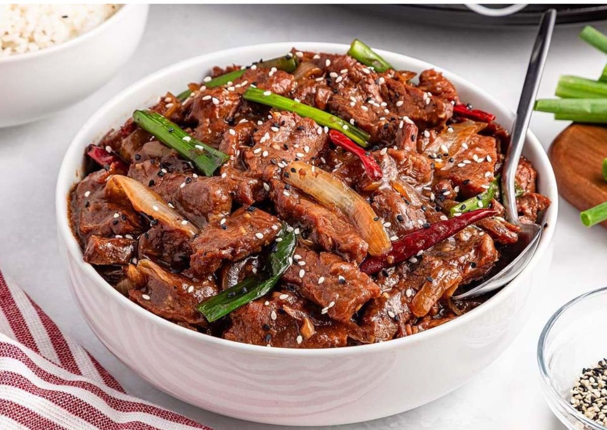 A bowl of stir-fried beef with green onions, red chilies, and sesame seeds, served with a spoon on a white surface.