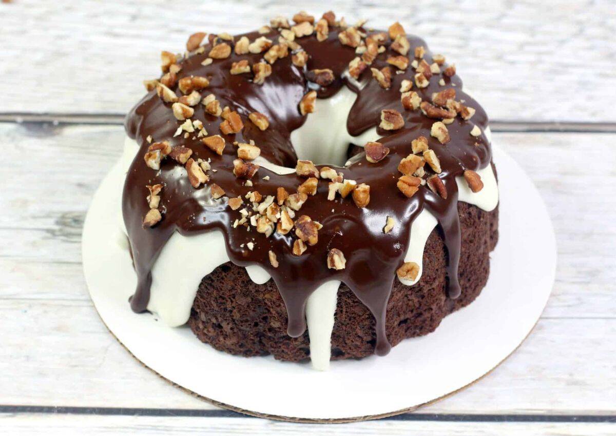 A chocolate bundt cake with white and dark chocolate glaze, topped with chopped nuts, sits on a white round base.