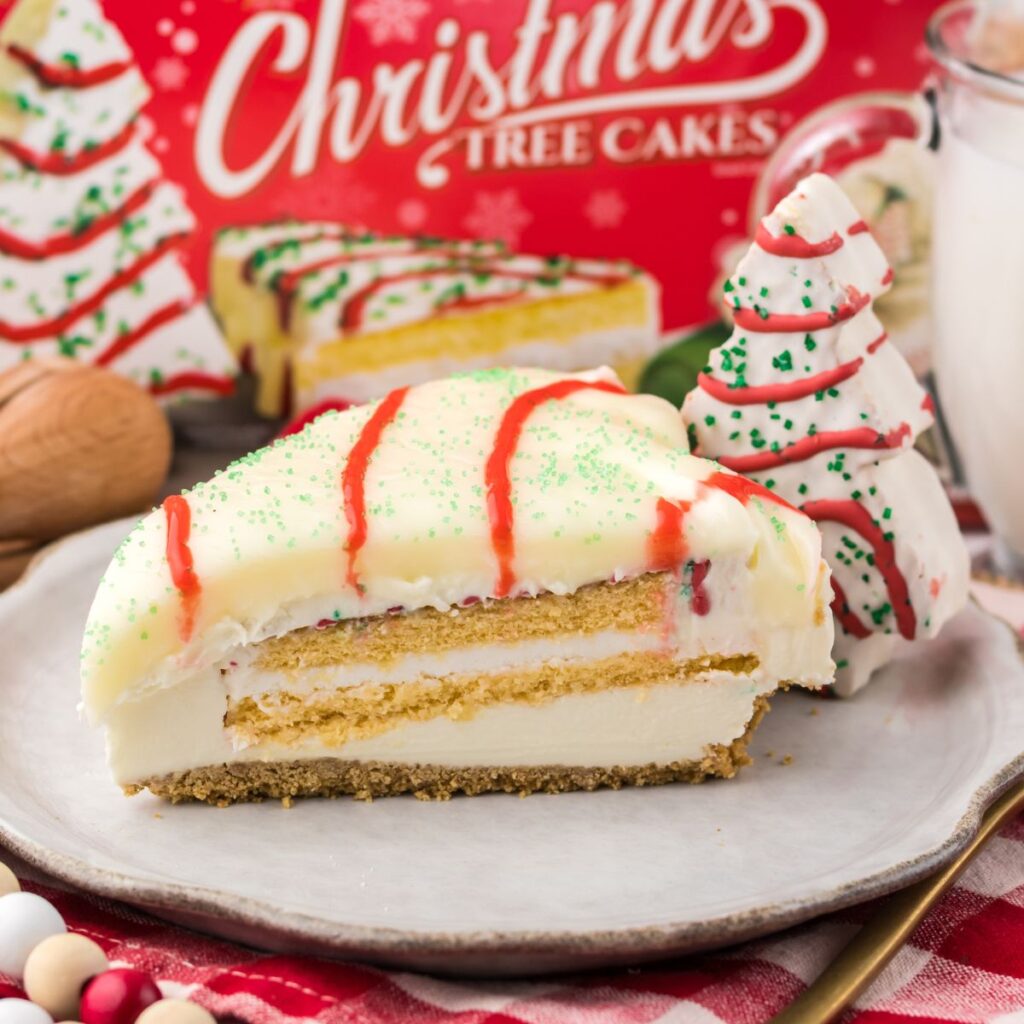 A slice of layered Christmas tree cake with white icing, green sprinkles, and red drizzle on a plate, with a tree-shaped cupcake and a glass of milk in the background.