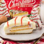 A slice of layered Christmas tree cake with white icing, green sprinkles, and red drizzle on a plate, with a tree-shaped cupcake and a glass of milk in the background.