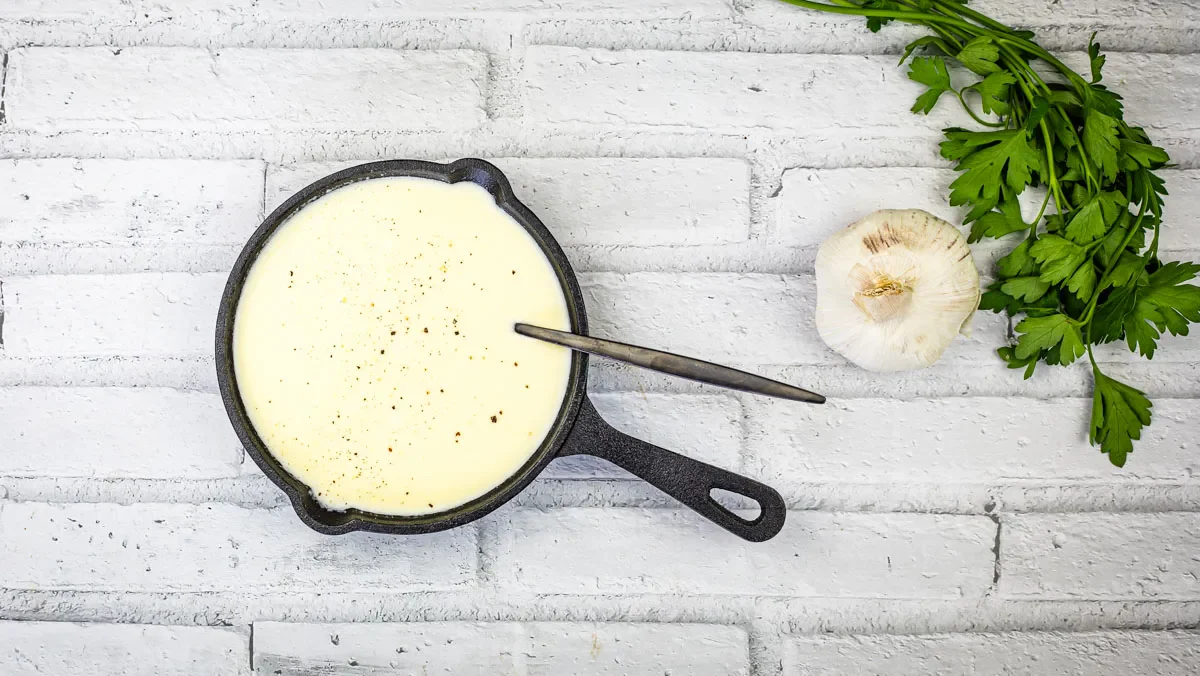 A small cast iron skillet filled with creamy white sauce, sprinkled with black pepper, sits on a white brick surface next to a garlic bulb and fresh parsley.