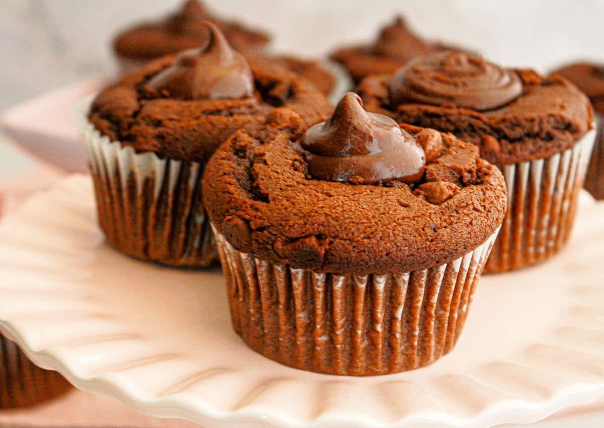 Three chocolate cupcakes with chocolate frosting swirl on top, displayed on a white scalloped cake stand.