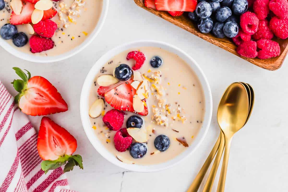 A bowl of cereal topped with sliced strawberries, blueberries, raspberries, and almond slivers, next to gold spoons and a dish of fresh berries on a white surface.