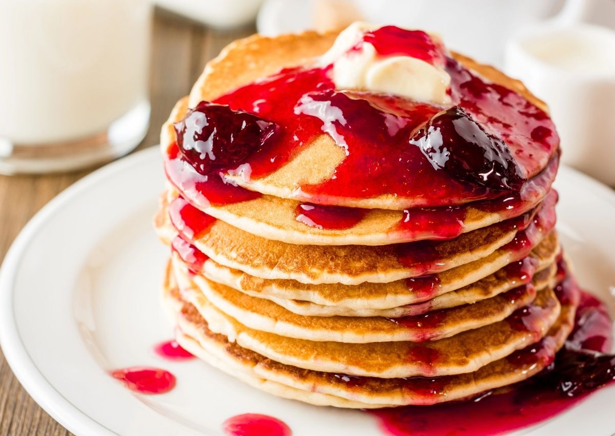 A stack of pancakes topped with a pat of butter and red berry syrup on a white plate.