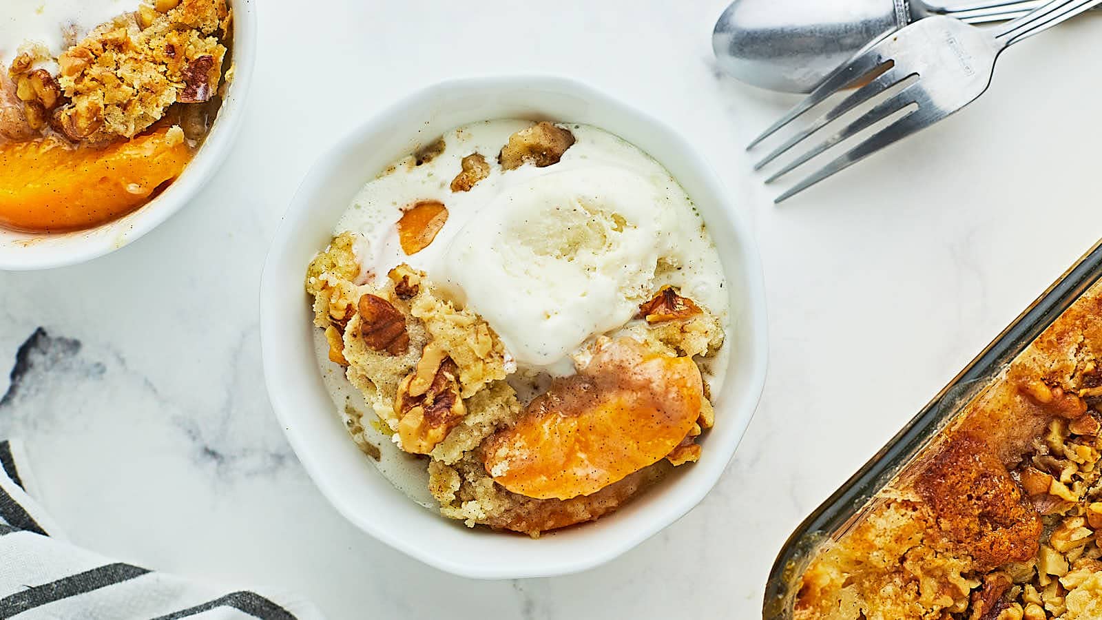 A bowl of peach cobbler topped with vanilla ice cream sits on a white surface, next to another serving and a baking dish. A fork and spoon are placed nearby.