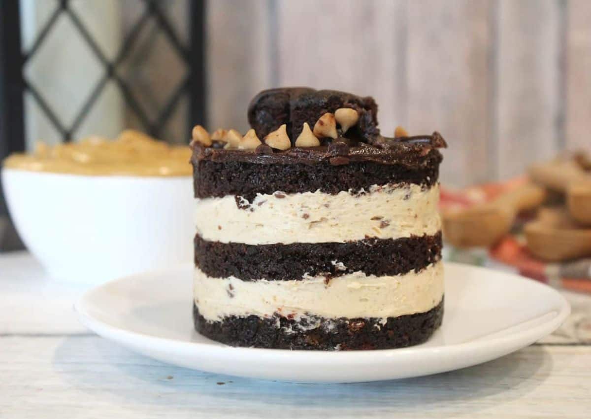 A layered chocolate and cream dessert topped with chocolate and nuts sits on a white plate; a bowl with yellow filling is in the background.