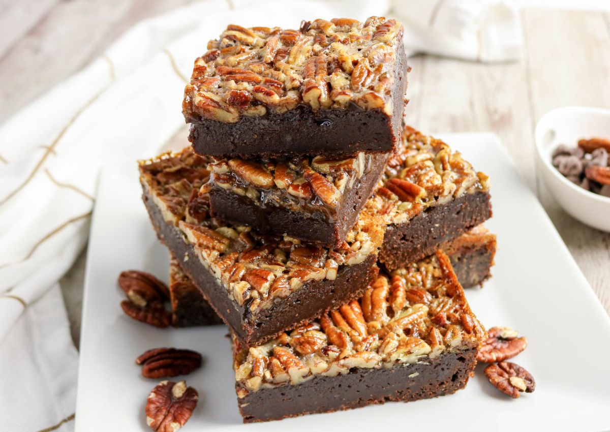 A stack of chocolate brownies topped with chopped pecans sits on a white plate, with additional pecans scattered around.