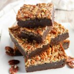 Four pecan-topped brownie bars are stacked on a white plate, with extra pecans scattered around them.