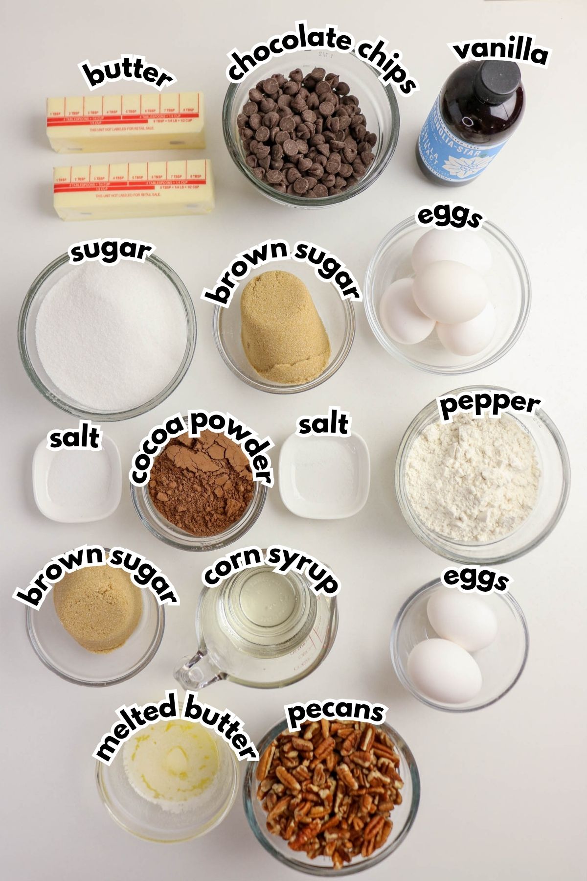 Top view of baking ingredients in bowls labeled: butter, chocolate chips, vanilla, sugar, brown sugar, eggs, cocoa powder, salt, pepper, corn syrup, melted butter, and pecans.