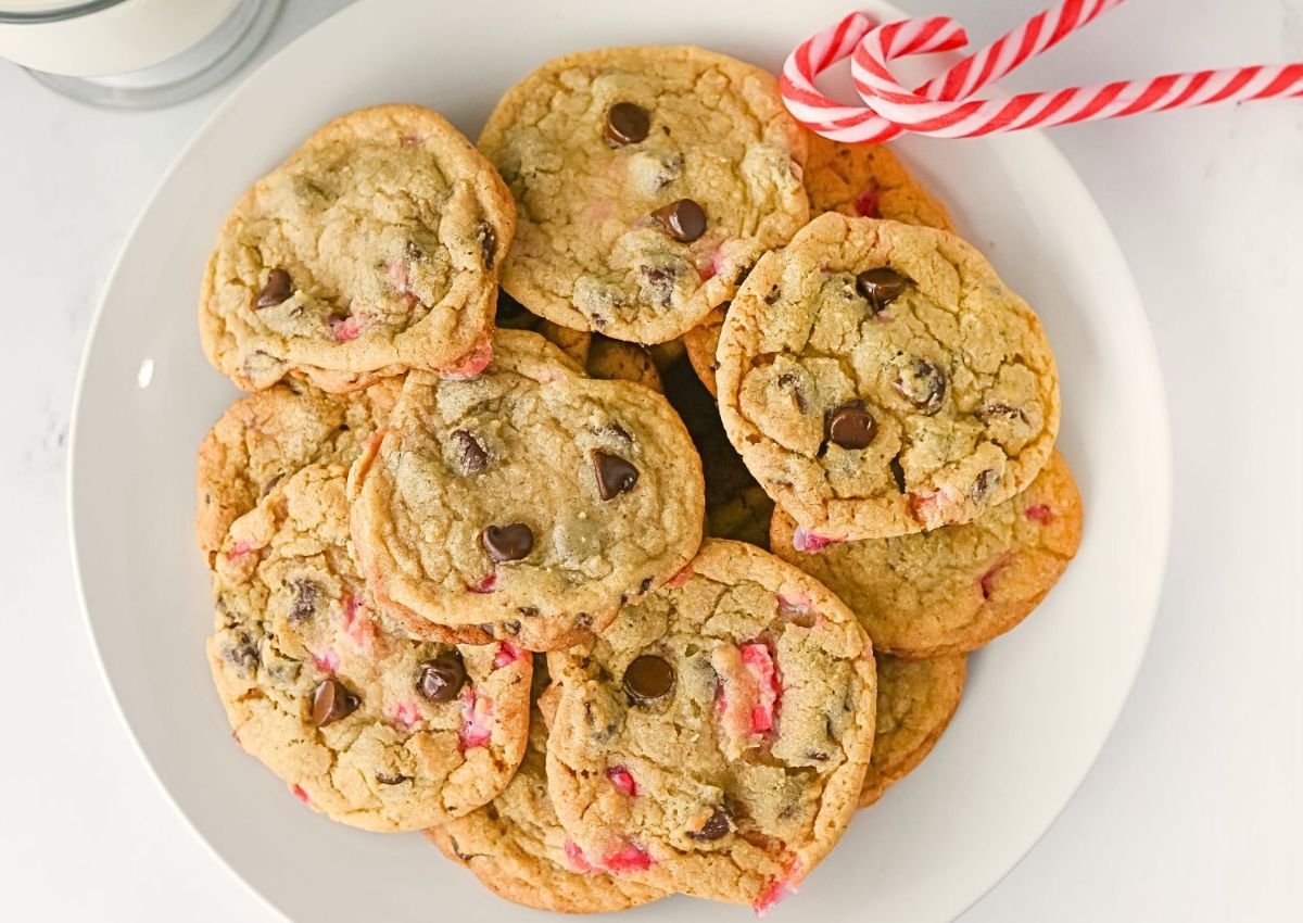 A white plate filled with chocolate chip cookies, some with visible red pieces, next to two candy canes and a glass of milk.