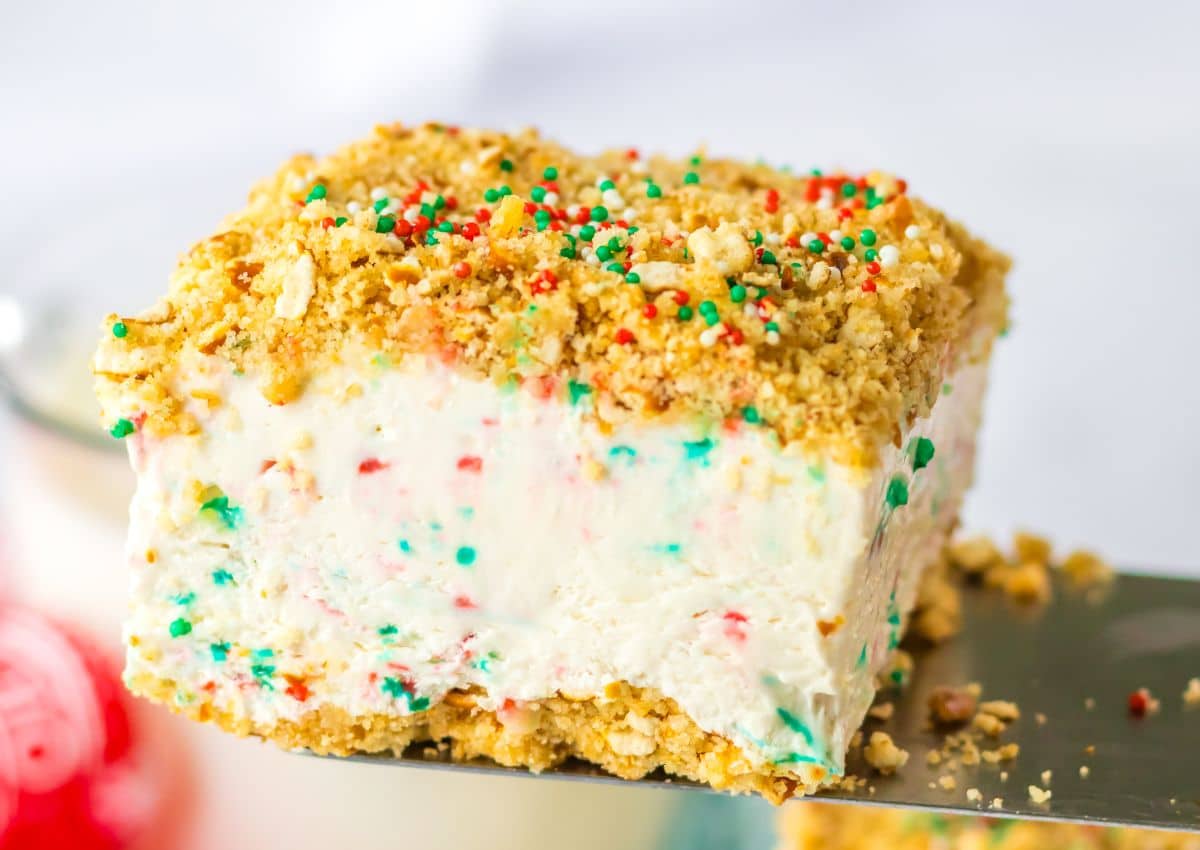 A slice of creamy dessert with a crumbly crust, colorful sprinkles mixed in and on top, served on a metal spatula.