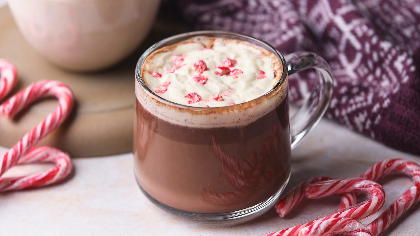 A clear glass mug of hot chocolate topped with whipped cream and crushed candy canes, surrounded by whole candy canes and a patterned purple cloth.