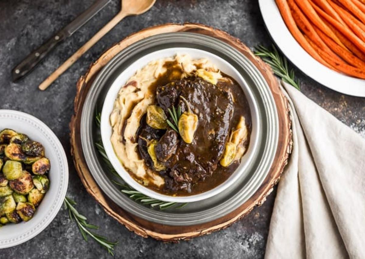 A bowl of mashed potatoes topped with beef in dark gravy and herbs, surrounded by side dishes of roasted Brussels sprouts and sliced carrots.