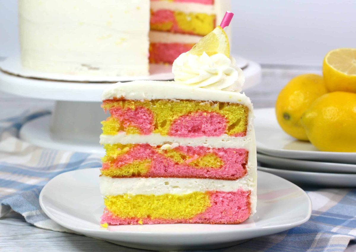 A slice of yellow and pink marbled cake with white frosting, topped with whipped cream and a lemon slice, sits on a plate; whole lemons and the rest of the cake are in the background.