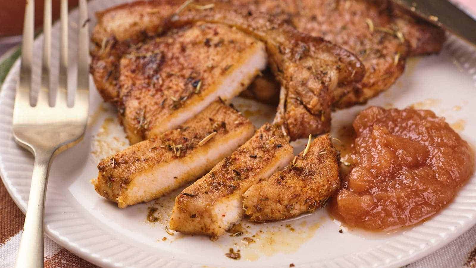 A plate with seasoned, cooked pork chops, some sliced, served alongside a portion of applesauce with a fork on the side.