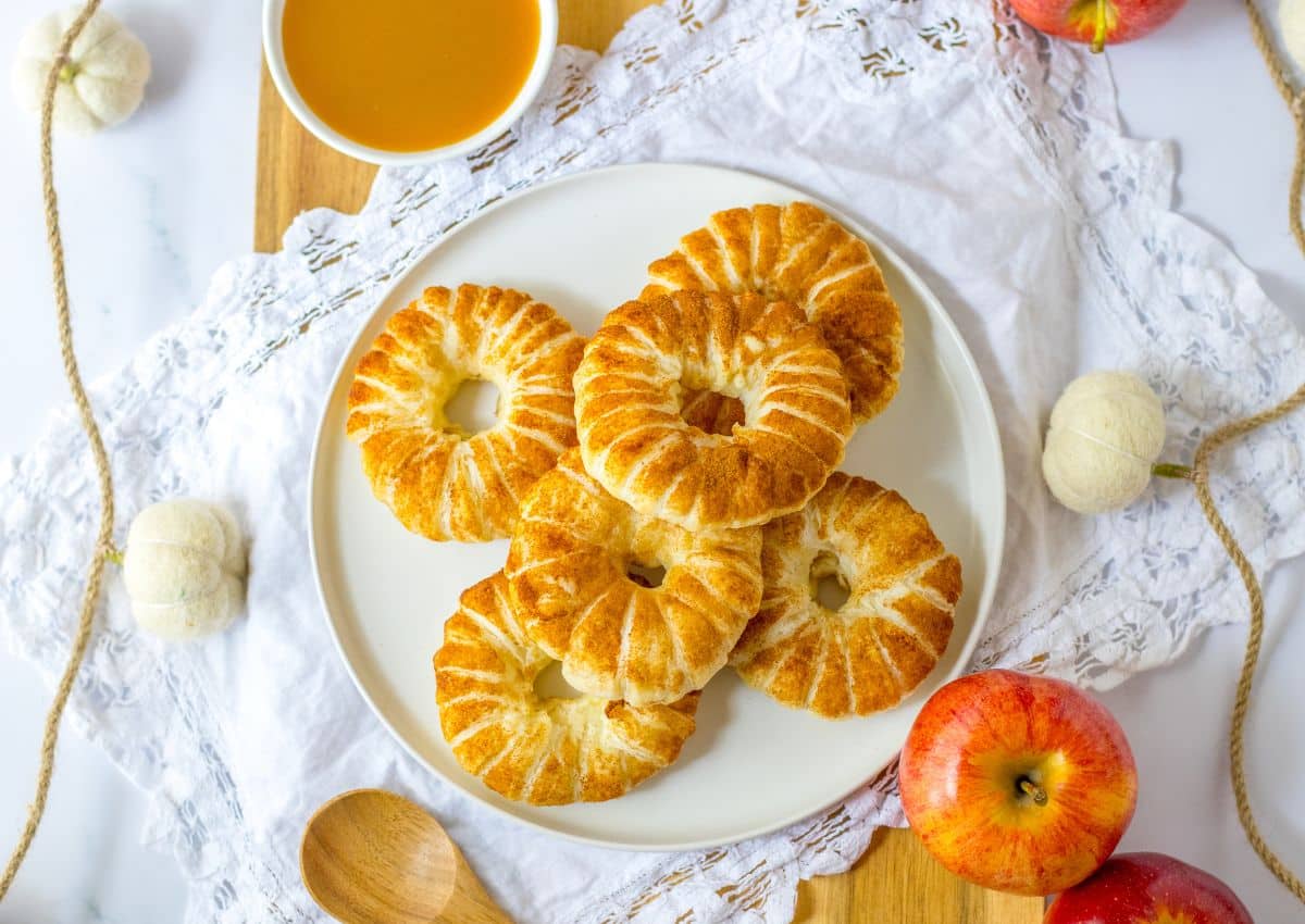 A plate of six round, glazed apple pastries sits on a white cloth, with apples, a small wooden spoon, and a bowl of caramel sauce nearby.