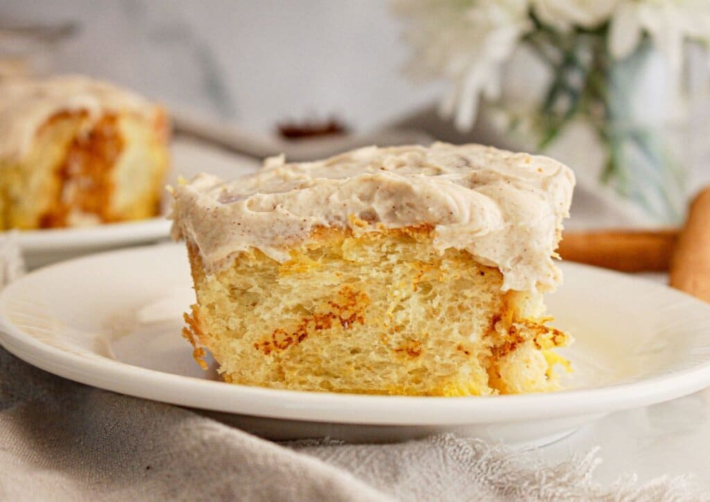 A close-up of a frosted cinnamon roll slice on a white plate, with another slice and blurred flowers in the background.