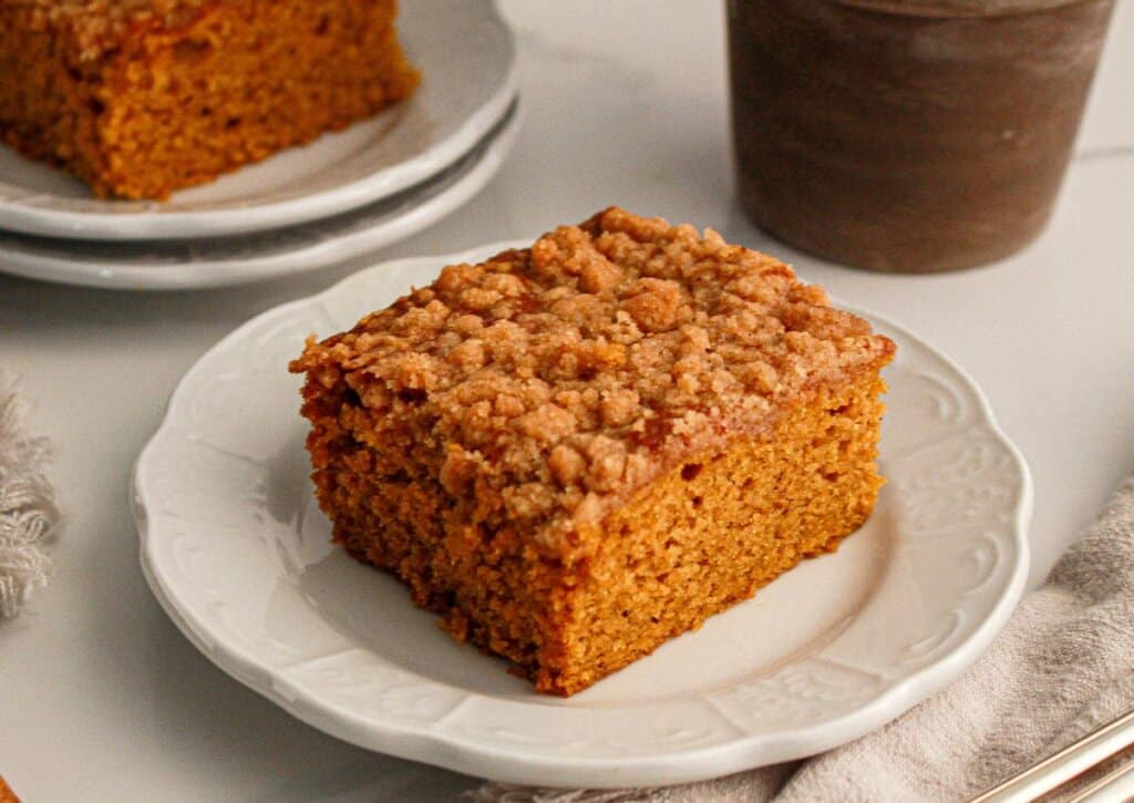 A slice of crumb-topped pumpkin coffee cake sits on a white plate, with another slice and a cup in the background.