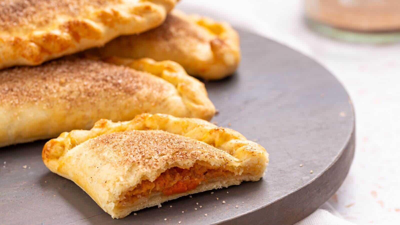 Three baked empanadas sit on a gray plate, with one empanada cut open to show a seasoned filling inside.
