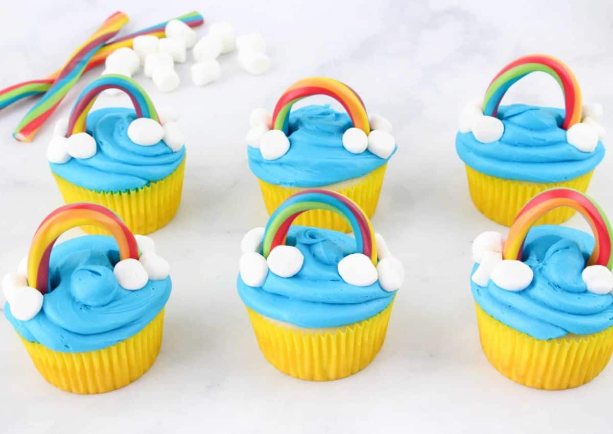 Six cupcakes with yellow wrappers, topped with blue frosting, rainbow candy arches, and small marshmallows on a white surface. Extra candies and marshmallows in the background.