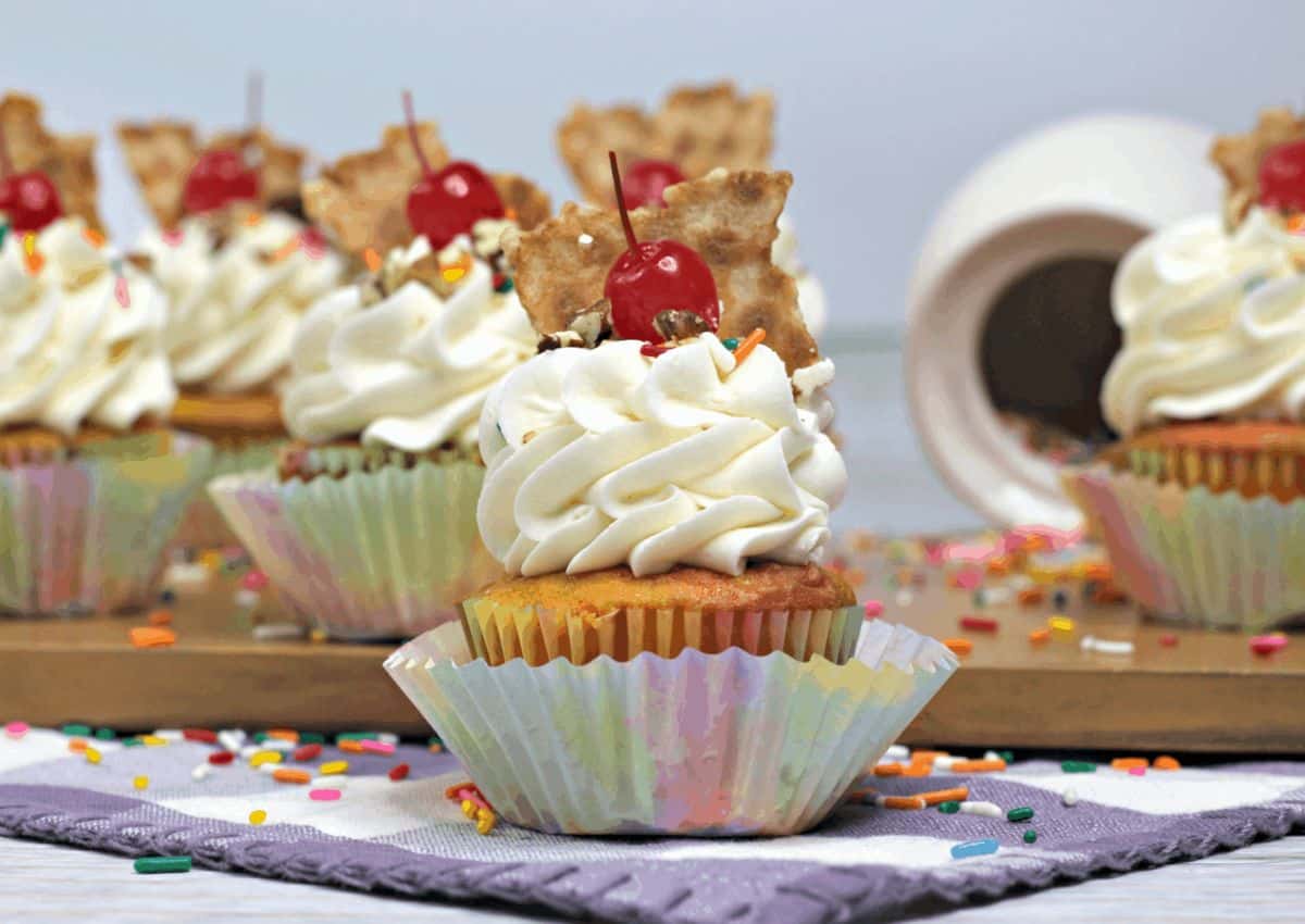 Cupcake with white frosting, a cherry, and a pastry piece on top, surrounded by other decorated cupcakes and scattered colorful sprinkles.