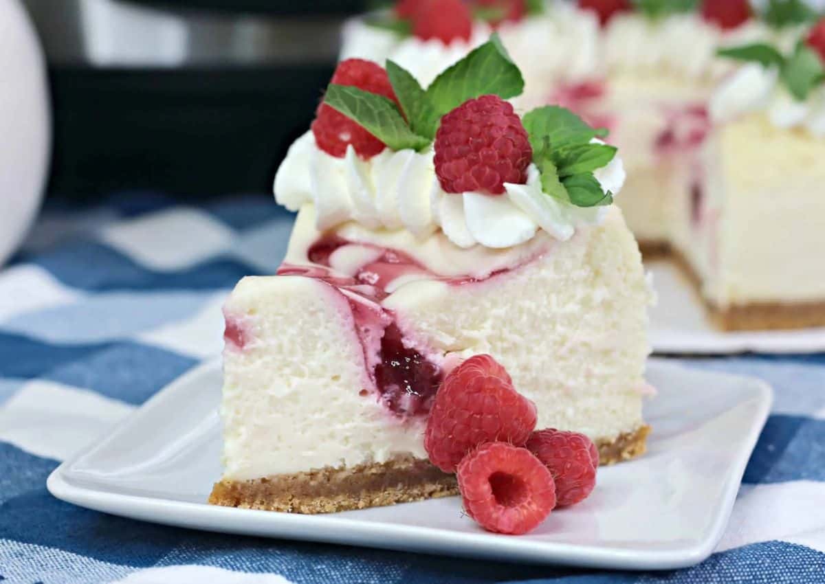 A slice of raspberry cheesecake with whipped cream, fresh raspberries, and mint leaves on a white plate.