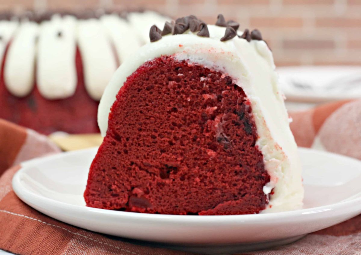 A slice of red velvet bundt cake with white cream cheese frosting and chocolate chips on a white plate.