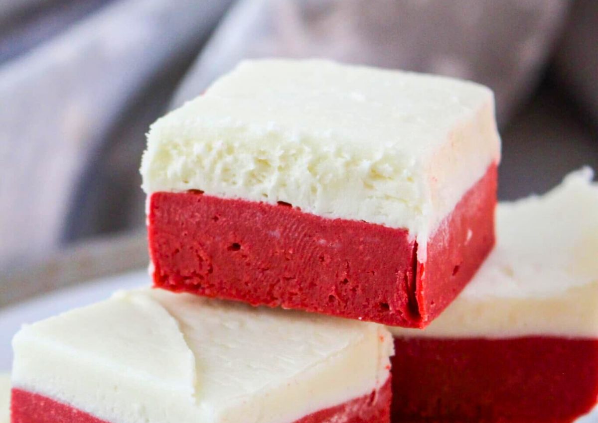 A close-up of two square pieces of red velvet fudge with a white cream cheese frosting layer on top.