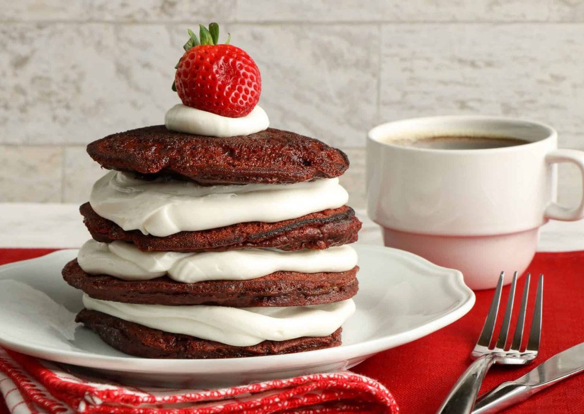 A stack of chocolate pancakes filled with cream, topped with a strawberry and whipped cream, sits on a white plate next to a cup of coffee, fork, and red napkin.