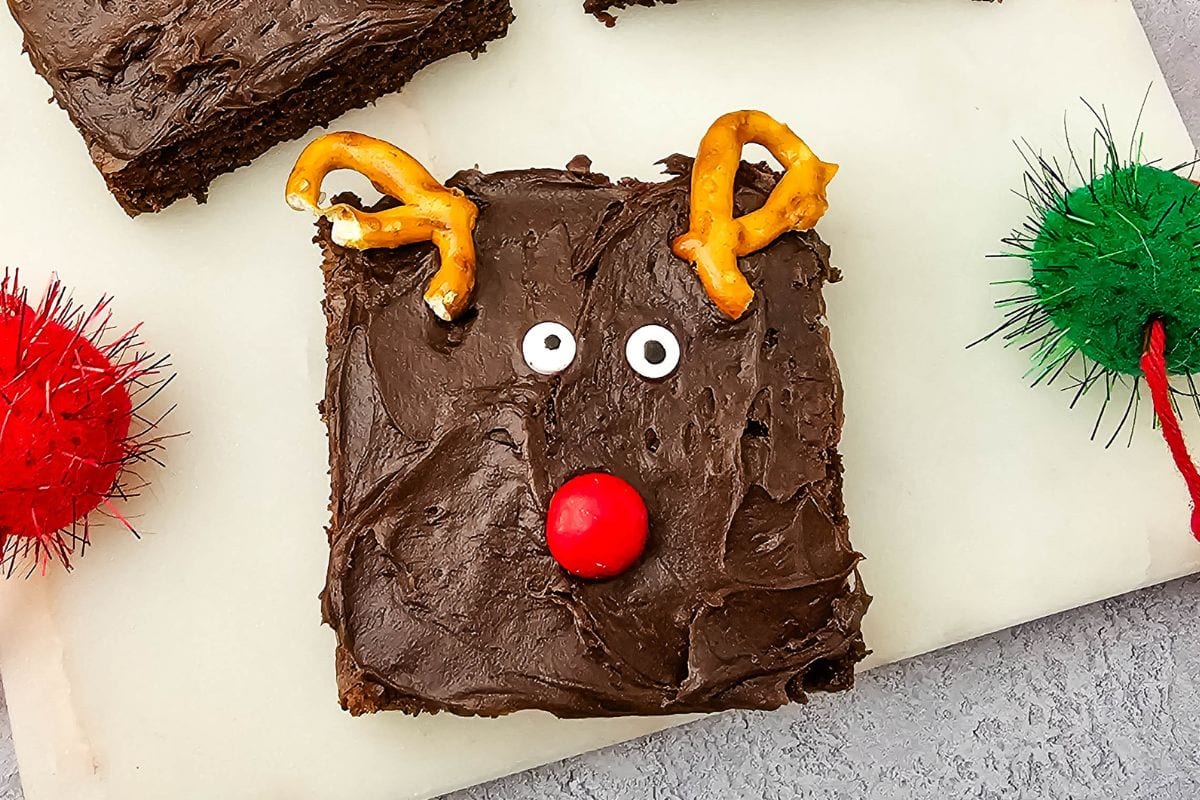 A brownie square with chocolate frosting, decorated as a reindeer using pretzels for antlers, candy eyes, and a red candy for the nose, placed on a white surface.