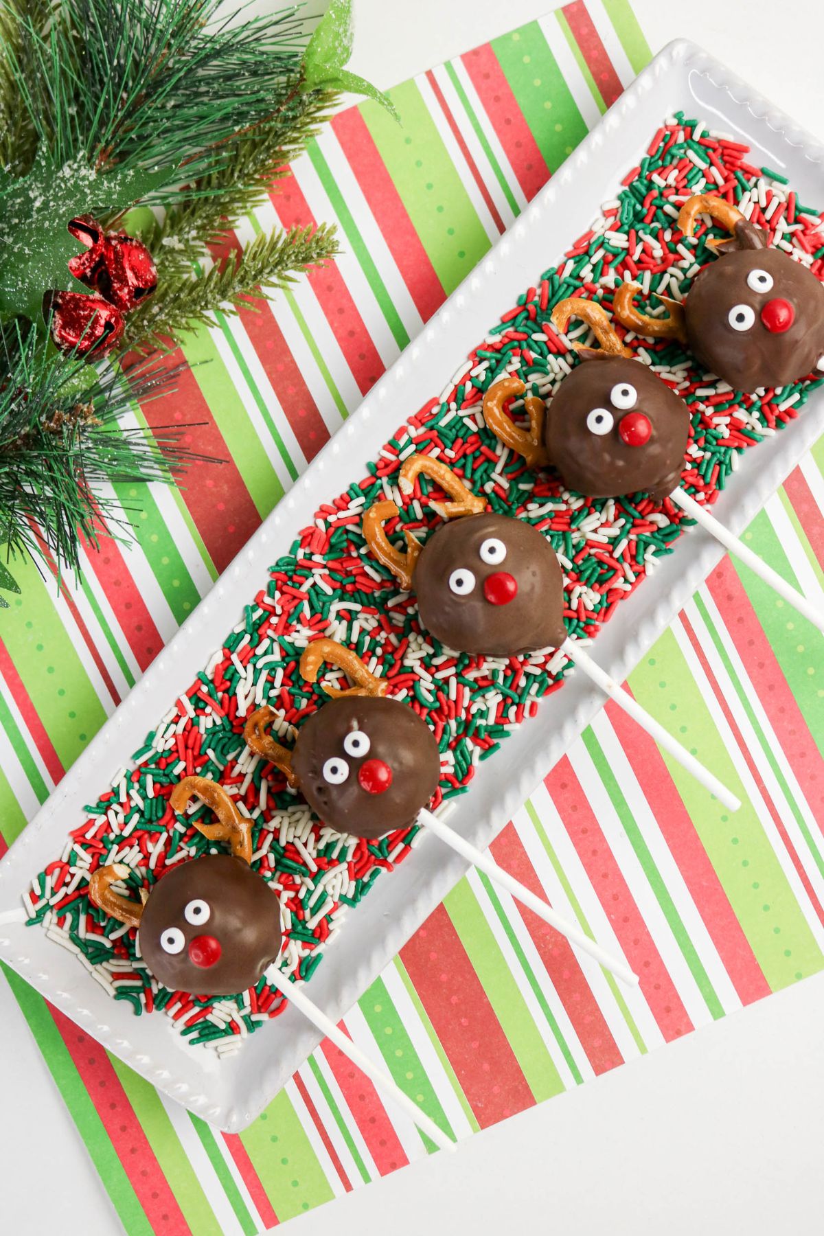 Five chocolate-covered reindeer cake pops with pretzel antlers and candy eyes are arranged on a white tray with holiday sprinkles, set on a red, green, and white striped placemat.