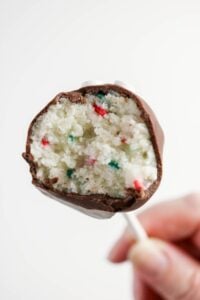 A close-up of a chocolate-covered cake pop with a bite taken out, revealing a white cake interior with red and green sprinkles.