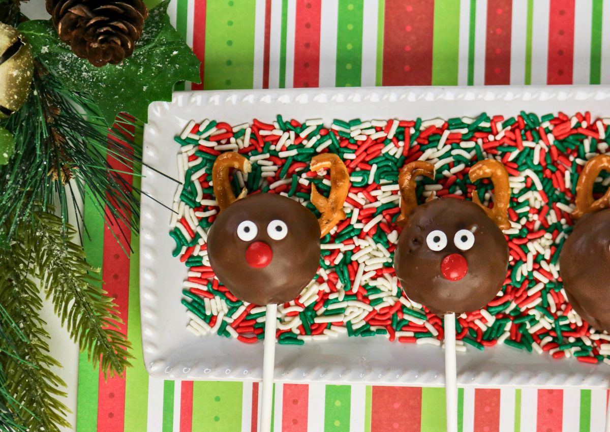 Chocolate reindeer cake pops with pretzel antlers and candy eyes on a plate covered in red, green, and white sprinkles, set on a festive striped background.