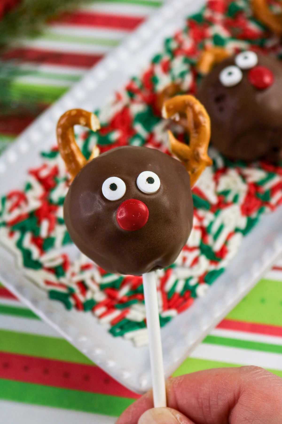 A hand holds a chocolate-covered cake pop decorated as a reindeer with candy eyes, a red candy nose, and pretzel antlers; more reindeer cake pops are on a tray in the background.