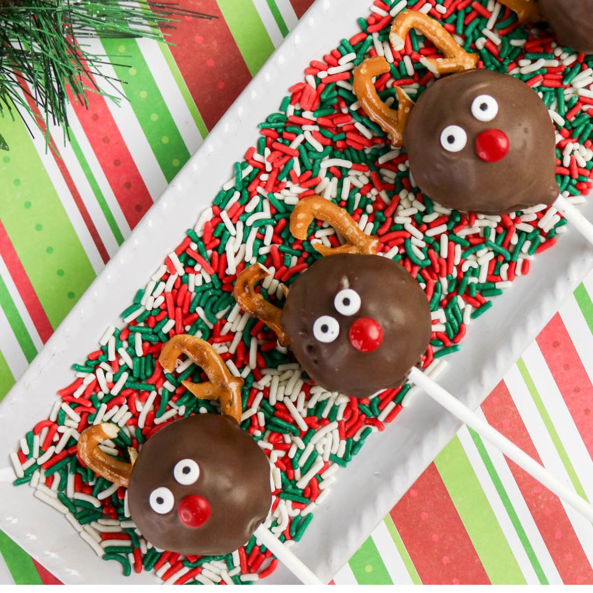 Three chocolate reindeer cake pops with candy eyes, red noses, and pretzel antlers are arranged on a platter with red, green, and white sprinkles.