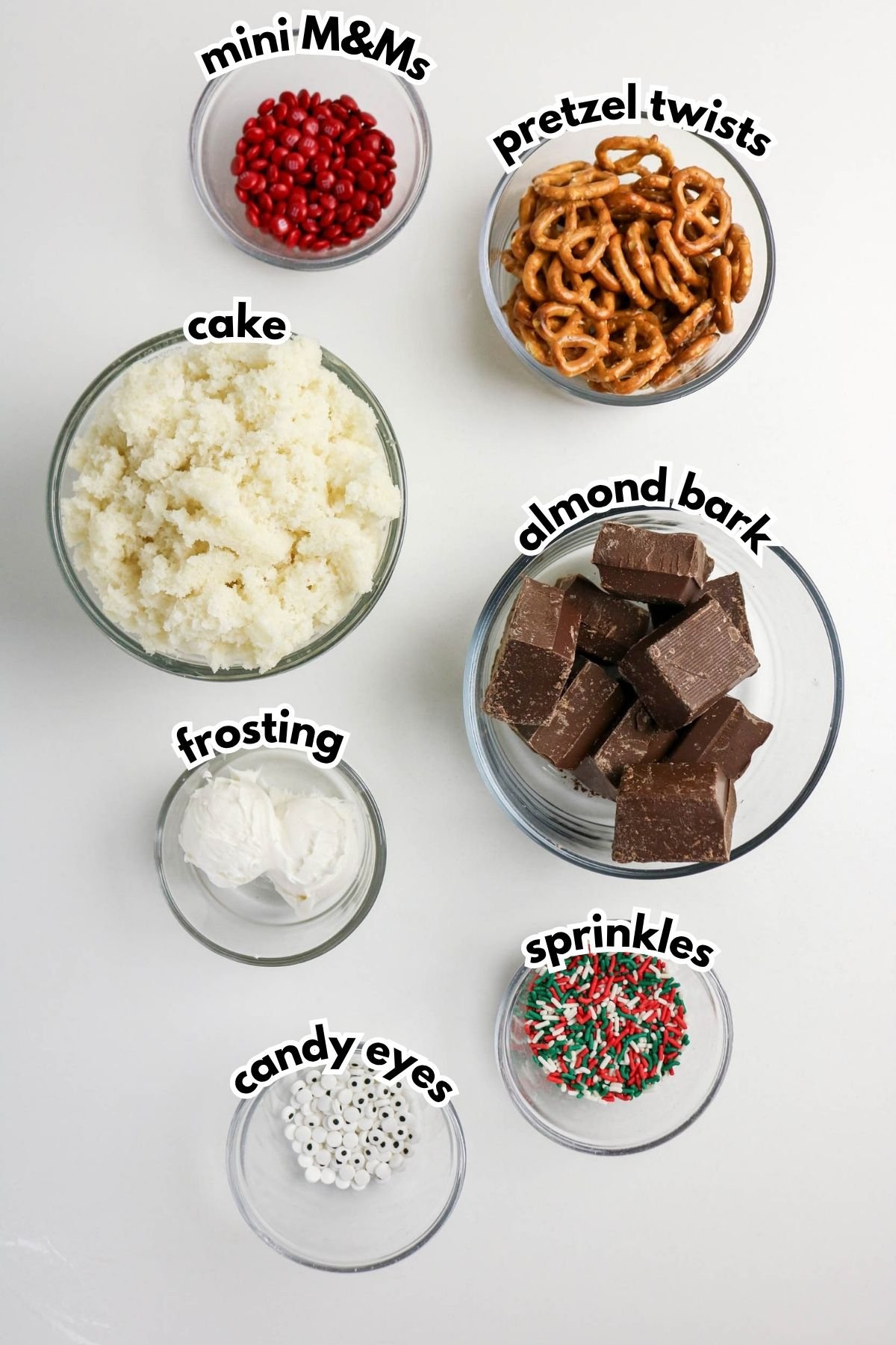 Seven bowls with labeled ingredients: mini M&Ms, pretzel twists, cake crumbs, almond bark, frosting, candy eyes, and sprinkles, arranged on a white surface.