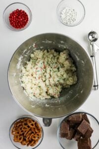 A mixing bowl with cookie dough and sprinkles is surrounded by bowls of red candies, candy eyes, pretzels, chocolate blocks, and a cookie scoop on a white surface.